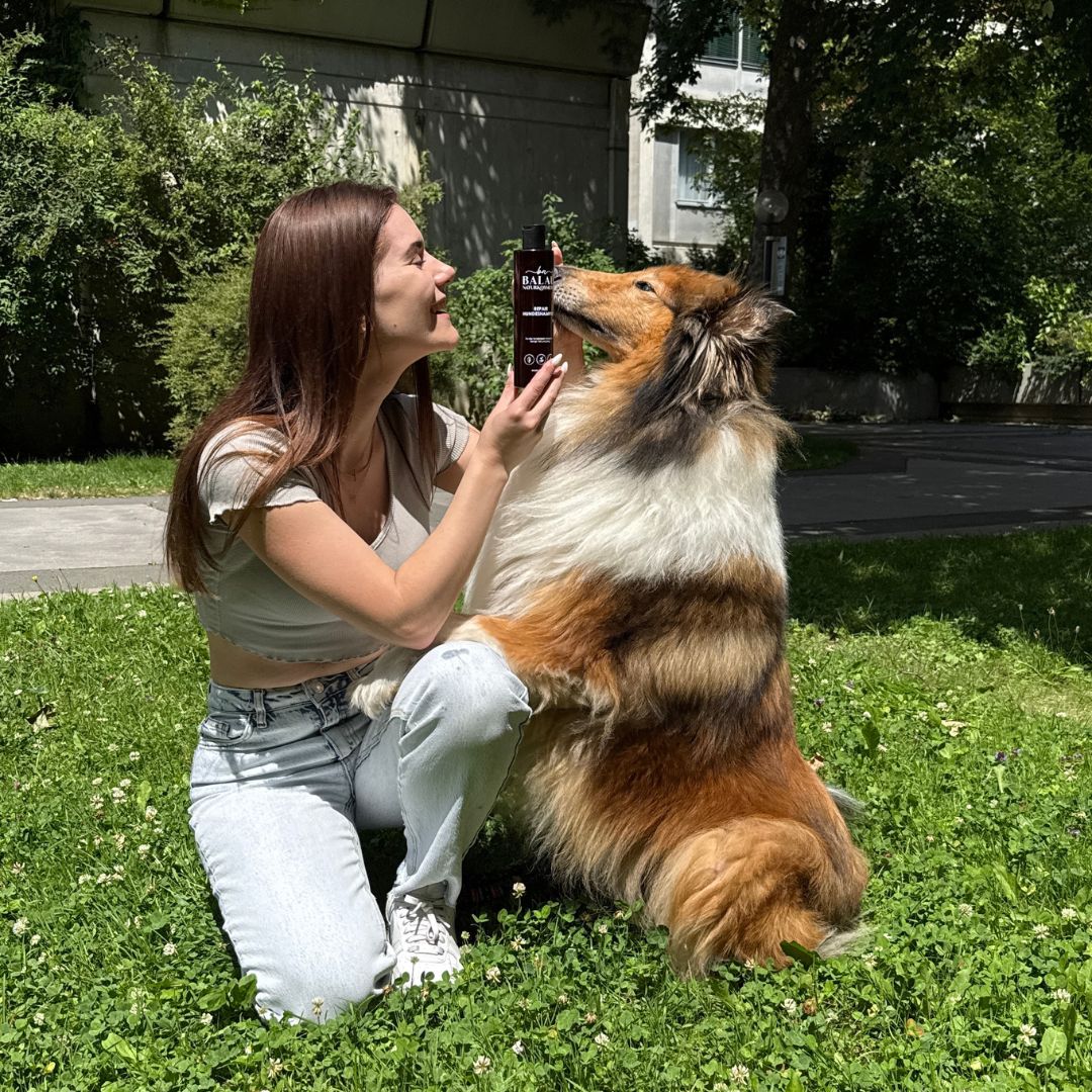 Frau sitzt mit Hund auf einer Wiese und zeigt das Balau Repair Hundeshampoo – natürliche Fellpflege für entspannte Momente